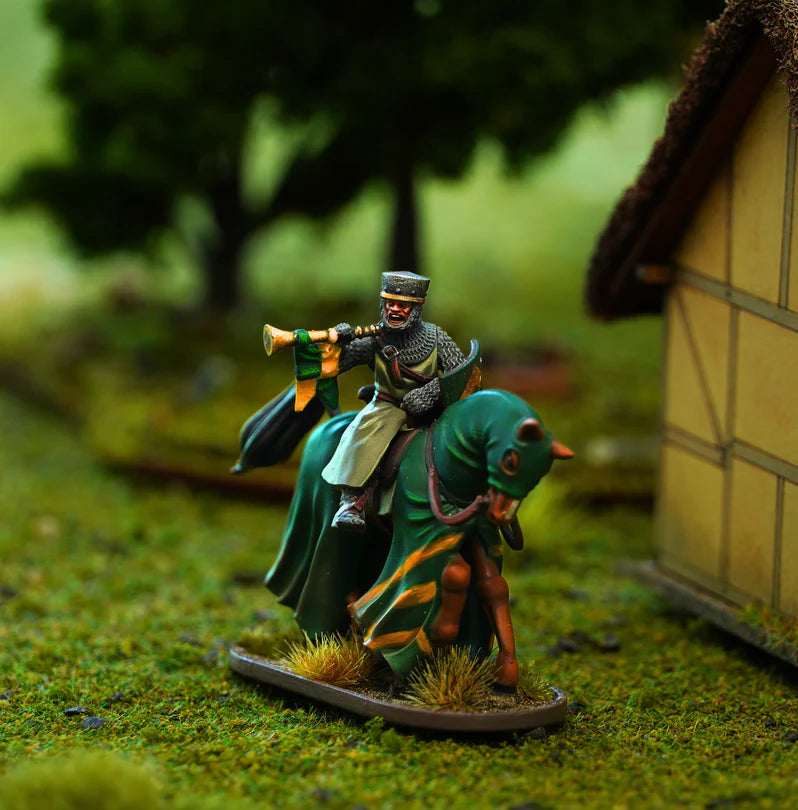 A painted tabletop miniature figurine of a knight in chainmail and helmet riding a green horse, holding a horn, set on grassy terrain with a blurred diorama background including a building and trees.