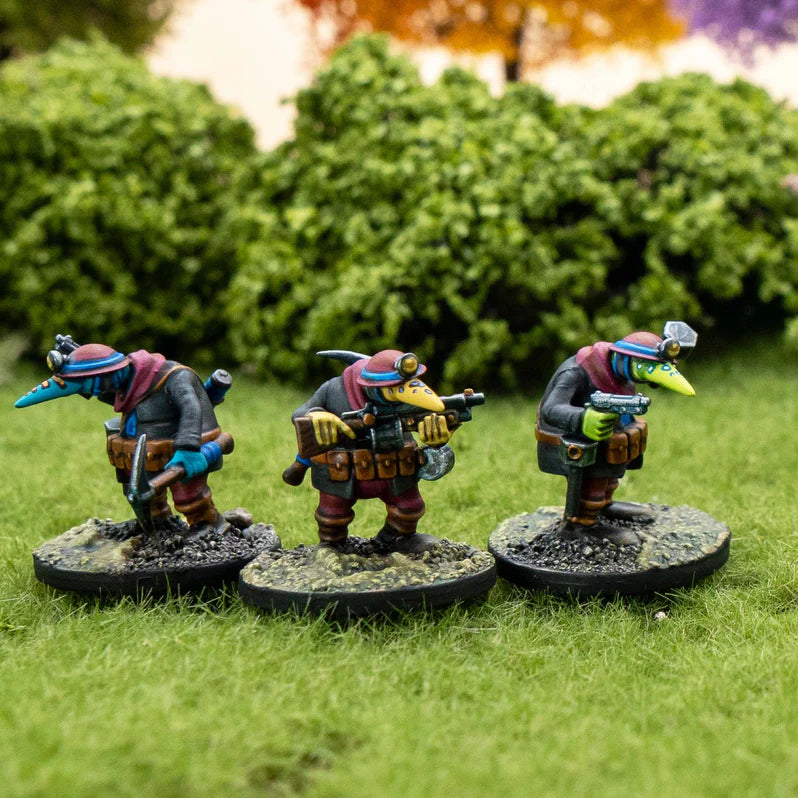 Three miniature figures with colorful outfits and weapons on grass with blurred greenery in the background